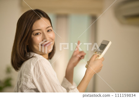 A middle-aged woman operates the remote control to turn on the air conditioner in the living room A middle-aged woman operates the remote control to turn on the air conditioner in the living room 111159834