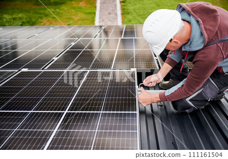 Man worker mounting photovoltaic solar panels on roof of house. Engineer in helmet installing solar module system with help of hex key. Concept of alternative, renewable energy. Man worker mounting photovoltaic solar panels on roof of house. Engineer in helmet installing solar module system with help of hex key. Concept of alternative, renewable energy. 111161504