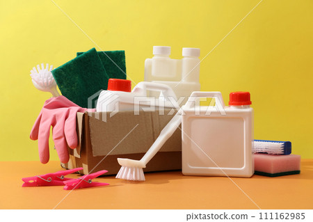 Front view of set of different detergents displayed on a yellow background. White plastic canisters without label decorated with brushes, sponges and rubber gloves on a carton box. Space for design 111162985