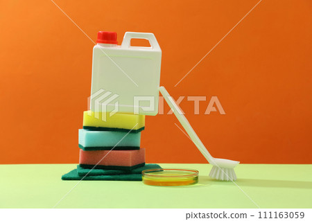 Creative background for advertising detergent products. Stack of sponges and detergent canister decorated with white brush and petri dish of yellow liquid on orange background. Blank space for design Creative background for advertising detergent products. Stack of sponges and detergent canister decorated with white brush and petri dish of yellow liquid on orange background. Blank space for design 111163059