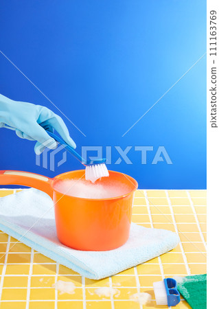 Front view of orange plastic water dipper container cleaning foam and wiper displayed on yellow ceramic background. Hand with rubber glove is holding a small brush. Advertising photo 111163769