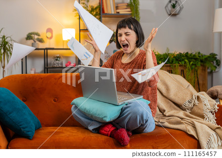 Exhausted woman freelancer with paperwork using laptop computer throwing documents in air at home 111165457