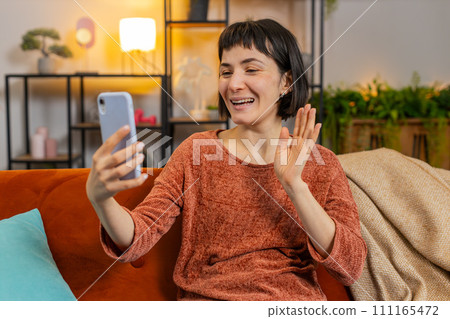Smiling young woman having conversation with friend on mobile smartphone sitting on sofa at home 111165472