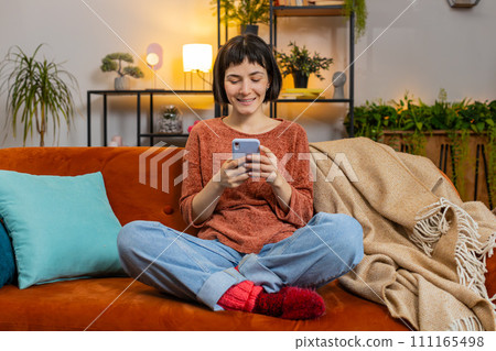 Happy woman thinking texting messaging on smartphone watching online movie sitting on home sofa 111165498