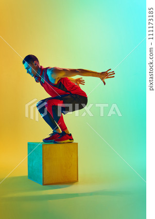 Full-length dynamic image of y9ung muscular man training, jumping on block, training against gradient blue yellow background in neon light Full-length dynamic image of y9ung muscular man training, jumping on block, training against gradient blue yellow background in neon light 111173185