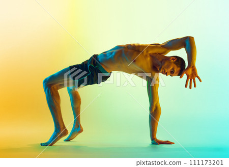 Intensive full-body training. Muscular athletic young shirtless man, with fit body doing workout exercises against gradient blue yellow background in neon light 111173201