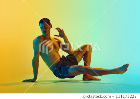 Young shirtless man with muscular, relief, strong body training, doing workout exercises against gradient blue yellow background in neon light Young shirtless man with muscular, relief, strong body training, doing workout exercises against gradient blue yellow background in neon light 111173222