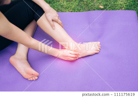 sportswoman holding her pain ankle after doing exercise on yoga mat in outdoor park. injury accident in exercise concept. sportswoman holding her pain ankle after doing exercise on yoga mat in outdoor park. injury accident in exercise concept. 111175619
