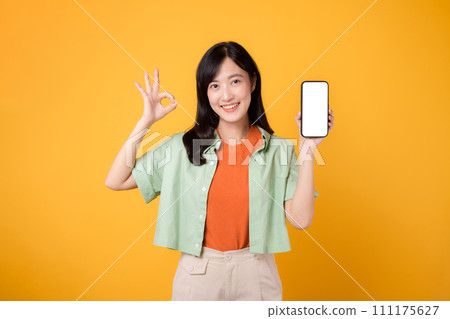 young Asian woman in her 30s, wearing orange shirt and green jumper, showcases smartphone screen display with an okay hand gesture on yellow studio background. New mobile application concept. 111175627