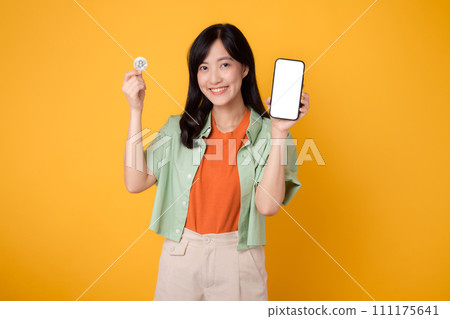 future finance with a cheerful young Asian woman in her 30s, donning orange shirt and green jumper, showcasing smartphone screen display and crypto currency coin on yellow background. 111175641