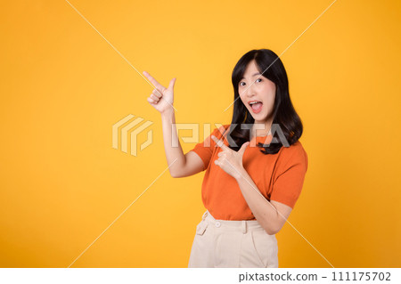 Spread happiness with a young Asian woman in her 30s, donning an orange shirt, pointing to free copy space on yellow background. recommending product concept. 111175702