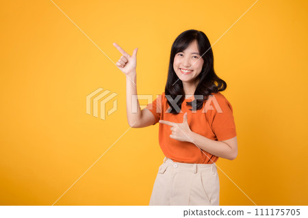 Radiate joy with a young Asian woman in her 30s, wearing an orange shirt, pointing to free copy space on yellow background. recommending product concept. 111175705