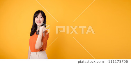 Feel the joy with a young Asian woman in her 30s, donning an orange shirt, pointing her thumb finger to free copy space on yellow background. product recommending concept. Feel the joy with a young Asian woman in her 30s, donning an orange shirt, pointing her thumb finger to free copy space on yellow background. product recommending concept. 111175706