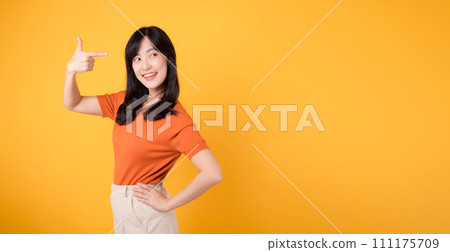 Feel the joy with a young Asian woman in her 30s, donning an orange shirt, pointing to free copy space on yellow background. recommending product concept. Feel the joy with a young Asian woman in her 30s, donning an orange shirt, pointing to free copy space on yellow background. recommending product concept. 111175709