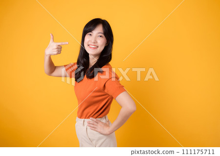 Cheerful young Asian woman in her 30s, wearing an orange shirt, points to free copy space with a happy face on yellow background. recommending product concept. Cheerful young Asian woman in her 30s, wearing an orange shirt, points to free copy space with a happy face on yellow background. recommending product concept. 111175711