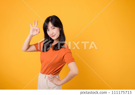 Embody joy with a young Asian woman in her 30s, donning an orange shirt, giving a okay hand gesture on yellow background. product recommending concept. Embody joy with a young Asian woman in her 30s, donning an orange shirt, giving a okay hand gesture on yellow background. product recommending concept. 111175715