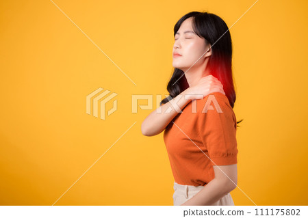 Compassionate Asian woman in her 30s, wearing an orange shirt, holds her painful neck on yellow background. Neck ache therapy medical office syndrome concept. 111175802
