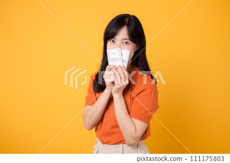 Smiling young modern 30s asian woman, holding cash money dollars, standing over yellow background 111175803