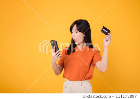 Satisfied Asian woman 30s in orange shirt, using smartphone and holding credit card on yellow background. Secure online payment. 111175897