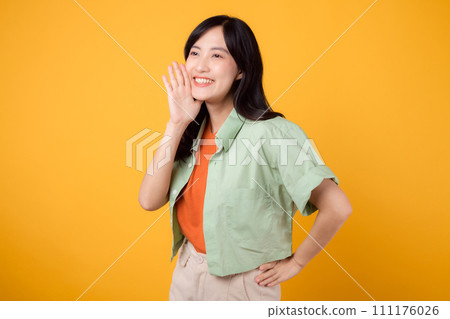 Energetic young Asian woman 30s wearing a green shirt on an orange background, enthusiastically shouting with excitement. Explore the concept of discount shopping promotion with this vibrant image. 111176026