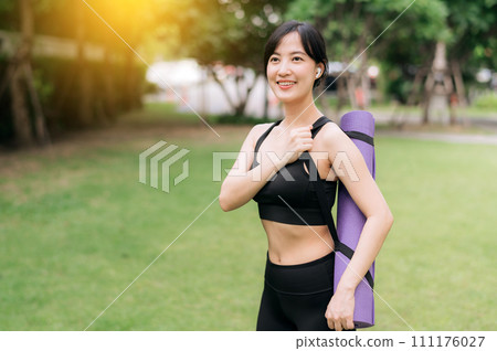 Yoga girl. Portrait of young asian slim and healthy girl doing workout in park, standing with rubber mat for exercises on green park at sunset, healthcare and well-being and yoga concept. 111176027