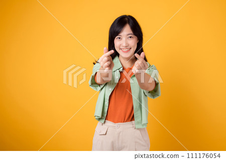 language of love with charming young Asian woman 30s. Her mini heart gesture and gentle smile convey affection dressed in orange shirt and green jumper, radiating warmth and joy. body language concept language of love with charming young Asian woman 30s. Her mini heart gesture and gentle smile convey affection dressed in orange shirt and green jumper, radiating warmth and joy. body language concept 111176054