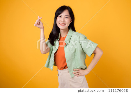 happiness with a young Asian woman in her 30s, dressed in an orange shirt and green jumper. Her mini heart gesture, hand on hip, and gentle smile convey a profound message through body language. happiness with a young Asian woman in her 30s, dressed in an orange shirt and green jumper. Her mini heart gesture, hand on hip, and gentle smile convey a profound message through body language. 111176058