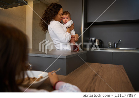 Mother hugs her baby boy, while older child daughter experiences jealousy looking at her mom carrying her little brother Mother hugs her baby boy, while older child daughter experiences jealousy looking at her mom carrying her little brother 111177887