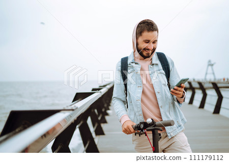 Young man with phone riding electric scooter on a pier near sea. Ecological transportation concept. 111179112