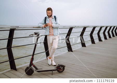 Young man with phone riding electric scooter on a pier near sea. Ecological transportation concept. Young man with phone riding electric scooter on a pier near sea. Ecological transportation concept. 111179119