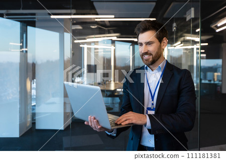Close-up photo of a smiling young businessman. a specialist programmer standing in a suit and with a badge in the office at a conference, holding and using a laptop. 111181381