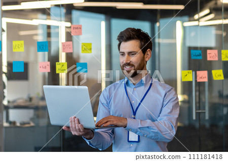 Smiling young man businessman, programmer and developer sitting inside a business center on the background of a glass board with notes and using a laptop in concentration. 111181418