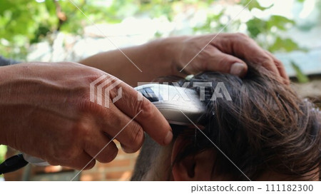 hands of a hairdresser cutting the back of a person's head with a clipper, close-up, shaving long dark hair from the back and top of the head with a trimmer with an attachment 111182300