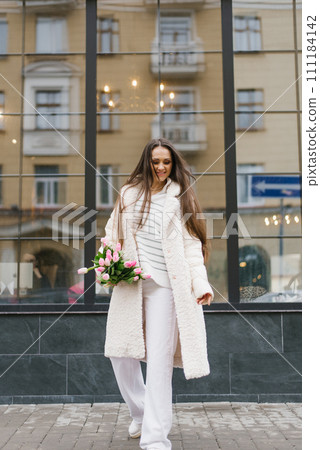Young beautiful woman in white coat with bouquet of white tulips flowers walking outdoors on the city street and smiles. Casual spring outfit 111184142