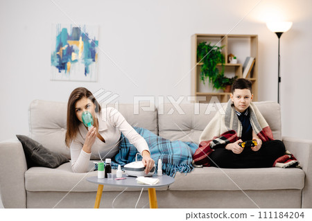 Young woman holding a mask nebulizer and inhaling fumes medication into lungs. Sick lady inhaling through inhaler mask at home and funny boy with gamepad playing video game at home near her 111184204