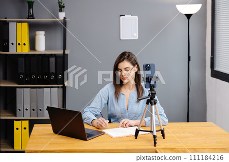 Videoconference And Training. Class of diverse students learning remotely online using web app. Female teacher providing lesson with laptop, recording video on cellphone on tripod 111184216