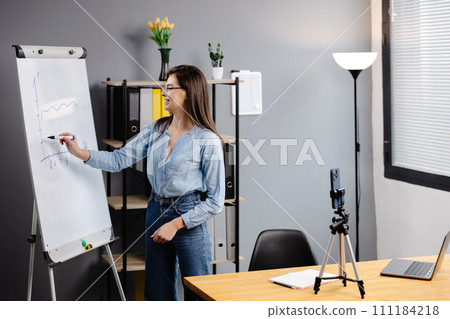 Young woman mentor recording presentation, webinar on smartphone standing on tripod, teacher coach explaining, holding online lesson, using phone webcam. 111184218