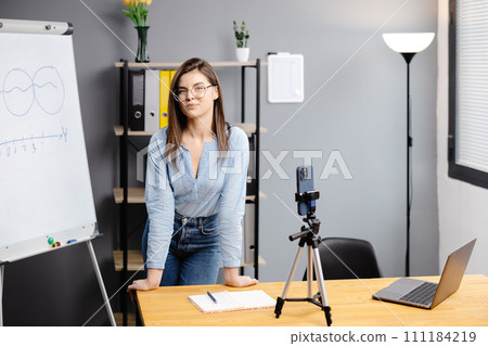 Young woman mentor recording presentation, webinar on smartphone standing on tripod, teacher coach explaining, holding online lesson, using phone webcam. 111184219