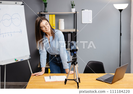 Positive blogger woman speaking at smartphone webcam on tripod, recording post for vlog. Online teacher, coach working, giving webinar at home. Presenter taking video on cellphone 111184220