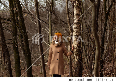 Woman in a brown coat between tree trunks in the forest Woman in a brown coat between tree trunks in the forest 111185493