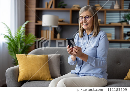 Senior gray-haired woman sitting joyfully on sofa at home, pensioner holding phone in hands, using app on smartphone, typing text message, call, and browsing social networks with a smile. Senior gray-haired woman sitting joyfully on sofa at home, pensioner holding phone in hands, using app on smartphone, typing text message, call, and browsing social networks with a smile. 111185509