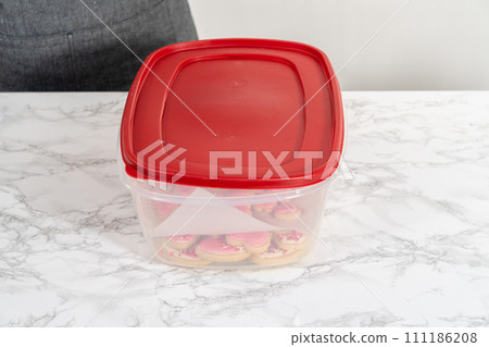 Storing heart-shaped sugar cookies with pink and white royal icing in a large plastic container. 111186208