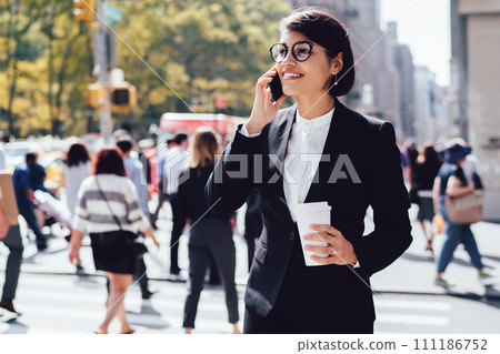 Positive corporate manager talking on phone during coffee break outdoors holding takeaway cup Positive corporate manager talking on phone during coffee break outdoors holding takeaway cup 111186752