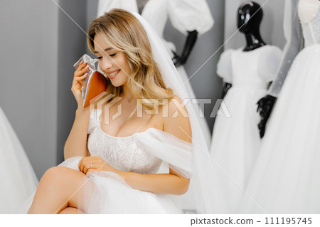 Bride-to-be wearing a wedding dress at a fitting room in a salon, choosing shoes for a wedding and having fun using a shoe as a phone Bride-to-be wearing a wedding dress at a fitting room in a salon, choosing shoes for a wedding and having fun using a shoe as a phone 111195745