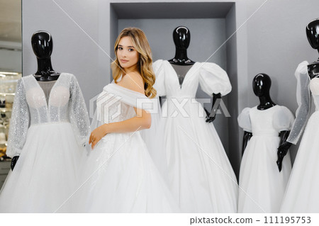 Pretty young woman is choosing a wedding dress in the shop. The bride-to-be is wearing a wedding dress for fitting Pretty young woman is choosing a wedding dress in the shop. The bride-to-be is wearing a wedding dress for fitting 111195753