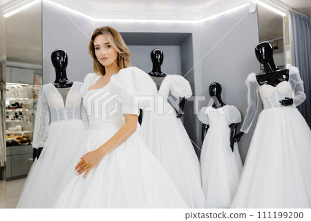 Pretty young woman is choosing a wedding dress in the shop. The bride-to-be is wearing a wedding dress for fitting 111199200