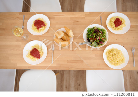 Top view of plates with appetizing spaghetti and classic tomato sauce, a wine glass of white wine and a bowl of fresh raw vegan salad on wooden table . Still life with traditional Italian food - pasta Top view of plates with appetizing spaghetti and classic tomato sauce, a wine glass of white wine and a bowl of fresh raw vegan salad on wooden table . Still life with traditional Italian food - pasta 111200811