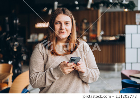 Young happy woman in glasses smiling and using smart phone in a cafe. Person using smartphone indoors. Online education, order, working or shopping concept Young happy woman in glasses smiling and using smart phone in a cafe. Person using smartphone indoors. Online education, order, working or shopping concept 111202778