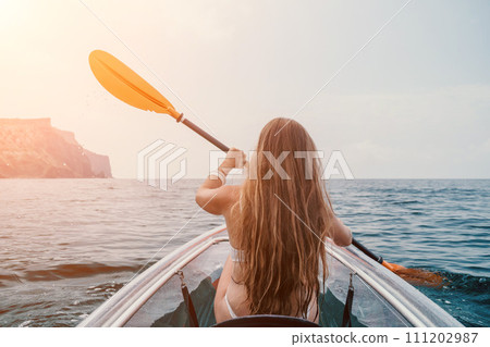 Woman in kayak back view. Happy young woman with long hair floating in transparent kayak on the crystal clear sea. Summer holiday vacation and cheerful female people relaxing having fun on the boat Woman in kayak back view. Happy young woman with long hair floating in transparent kayak on the crystal clear sea. Summer holiday vacation and cheerful female people relaxing having fun on the boat 111202987