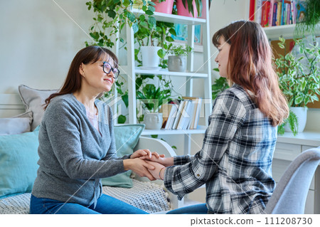Talking mother and teenage daughter sitting together on couch at home, smiling holding hands Talking mother and teenage daughter sitting together on couch at home, smiling holding hands 111208730